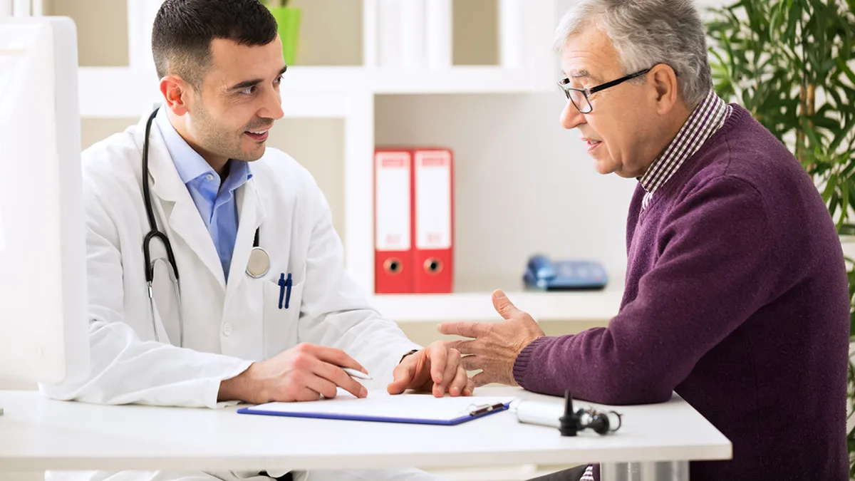 A doctor in a white coat talking across a desk with an older man in a consulting room